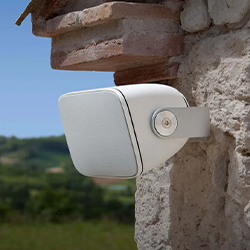 Security camera mounted on a stone wall, facing outward. Bright, sunny day with a blurred landscape of greenery and hills in the background.