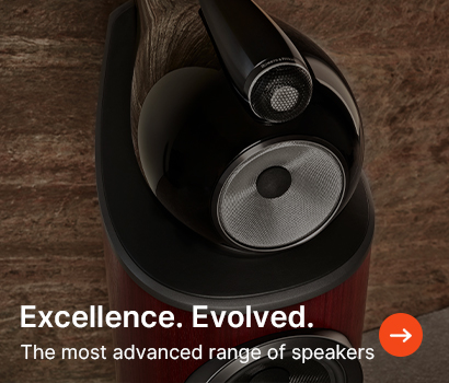 A sleek, modern speaker with wood accents on a textured background. Text reads "Excellence. Evolved. The most advanced range of speakers." Orange arrow icon.