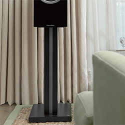A sleek black speaker on a stand is positioned in a room with beige curtains and a light green sofa, conveying a modern and serene ambiance.