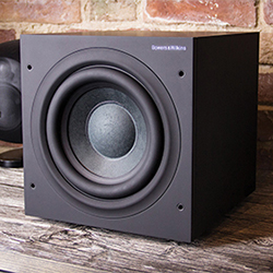A sleek black subwoofer sits on a wooden surface against a rustic brick wall, conveying a modern and stylish audio equipment setup.