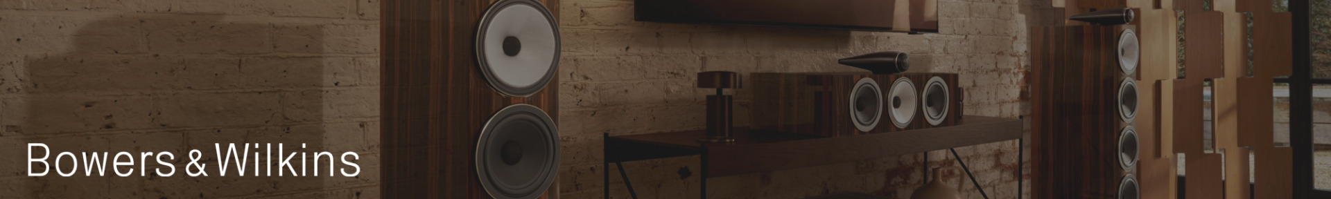 Modern audio setup with wooden speakers and a soundbar in a cozy room with brick walls. "Bowers & Wilkins" text on the left. Warm, sophisticated ambiance.