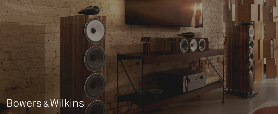 Modern audio setup with wooden speakers and a soundbar in a cozy room with brick walls. "Bowers & Wilkins" text on the left. Warm, sophisticated ambiance.