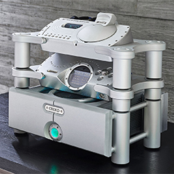 Futuristic audio equipment with a silver, modular design sits on a dark surface. Features include control dials and a glowing green button, conveying advanced technology.