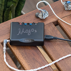 A compact black audio device labelled "Mojo 2" with colourful buttons is connected to earphones on a wooden table. 