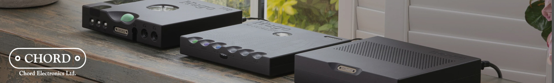Three sleek black audio devices from Chord Electronics are placed on a wooden table by a window. The scene is calm and modern, highlighting premium design.