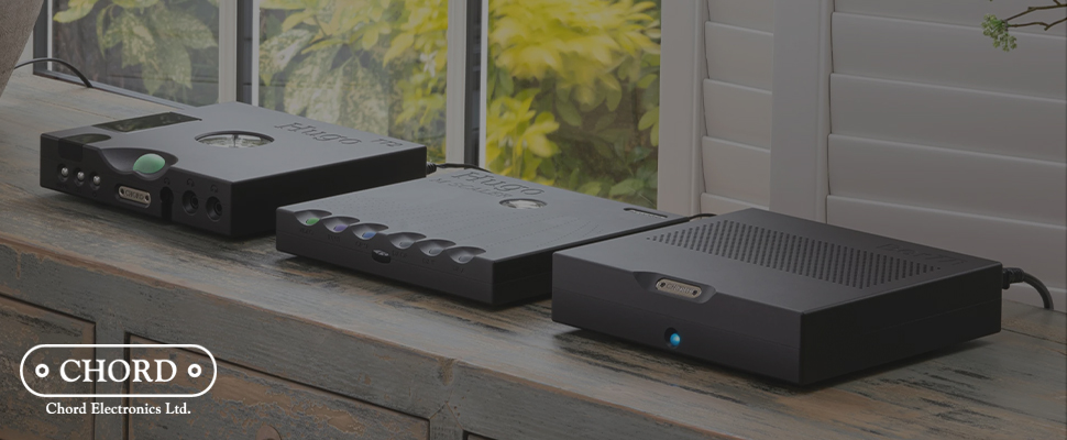 Three sleek black audio devices from Chord Electronics are placed on a wooden table by a window. The scene is calm and modern, highlighting premium design.