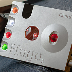 A metallic device with colourful buttons, labelled "Hugo 2" and "Chord," featuring glowing red, orange, white, and green elements, resting on denim fabric.
