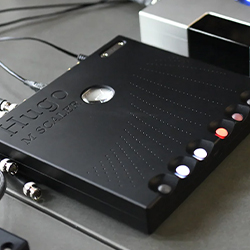 A black audio device with "Hugo" and "M Scaler" embossed on top. It features colourful LED buttons and cables connected, set on a grey surface.