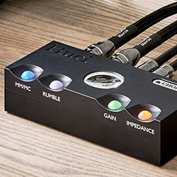 Black audio device with labels "MM/MC," "RUMBLE," "GAIN," and "IMPEDANCE" on a wooden surface. Cables connect at the back.