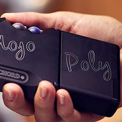 A hand holds a compact black audio device labelled "Mojo Poly" with rounded buttons. The background is softly blurred.