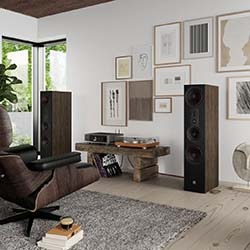 Modern living room with large speakers, turntable, and framed art on white walls. Brown chair on a grey rug conveys a cosy, stylish atmosphere.