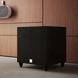 A sleek black subwoofer with a small logo sits on a modern, light-panelled floor.
