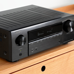 A black audio receiver with dials and buttons sits on a wooden surface. The digital display shows "Dolby Audio," conveying a modern, sleek design.