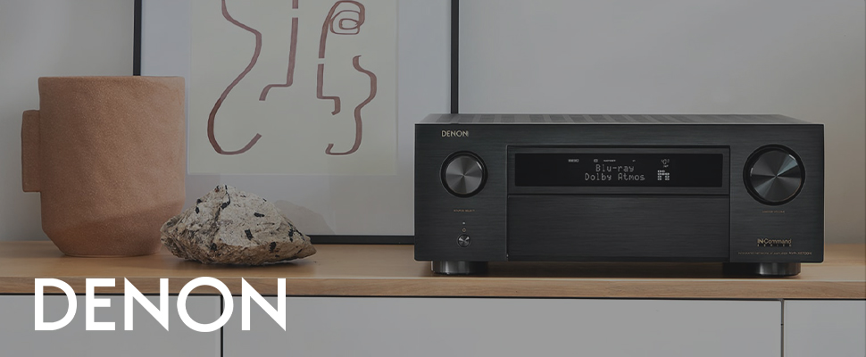 Denon stereo receiver on a wooden shelf with decor. Display shows “Blu-ray, Dolby Atmos.” Aesthetic setup with books and abstract art.