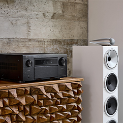 A sleek audio receiver on a textured wooden cabinet beside a modern white speaker with multiple drivers, set against a concrete wall.
