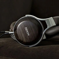 Sleek Denon headphones with a wood finish rest on a dark brown sofa, emitting a modern, warm vibe. The setting suggests a cosy listening atmosphere.