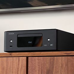 A sleek black audio receiver sits on a wooden cabinet, displaying "HDMI" on its front panel. The room is modern and minimalistic.