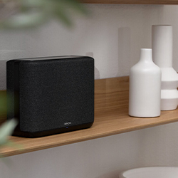 A sleek, black wireless speaker sits on a wooden shelf next to minimalist white vases, set against a soft, neutral wall, conveying a modern and cosy ambiance.