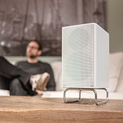 A sleek white speaker on a wooden table in focus, with a blurred background showing a person lounging on a sofa, creating a relaxed, modern ambiance.