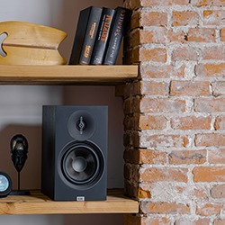 A black speaker is placed on a wooden shelf against a brick wall, next to a small mic and dark books, creating a modern, cosy atmosphere.
