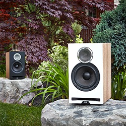 Two modern speakers placed on stone platforms in a lush garden setting, surrounded by vibrant green plants and textured tree foliage.
