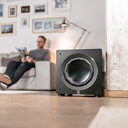 A man sits on a sofa reading in a cosy living room. A large black speaker is in the foreground. The wall is adorned with framed photos and art.