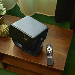 A black projector and a remote sit on a wooden table against a green backdrop, nearby a potted plant and sheer curtains, creating a cosy ambiance.