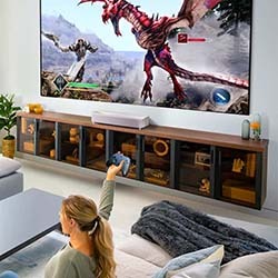 A person sitting on a couch plays a video game on a large TV, battling a red dragon. The room is modern with a sleek entertainment unit.