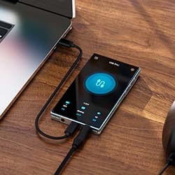 A sleek, modern digital audio player connected to a laptop on a wooden desk. The device screen displays audio settings, exuding a technological and efficient vibe.