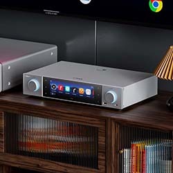 A sleek audio receiver with a digital display sits on a wooden shelf, showing various app icons. Books and a gold lamp are nearby, creating a cosy ambiance.