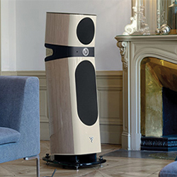 A modern beige speaker with black accents stands in a stylish living room. Nearby is a blue sofa and an ornate fireplace, creating an elegant atmosphere.