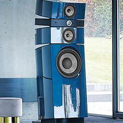 Sleek, modern blue floor-standing speaker with multiple drivers, set in a minimalist room with a large window and soft natural light.