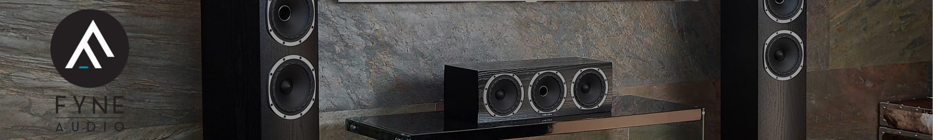 Modern home audio system with two tall black speakers and a central speaker on a sleek glass stand. Background features a stone-textured wall. Fyne Audio logo on the left conveys an elegant, high-end feel.