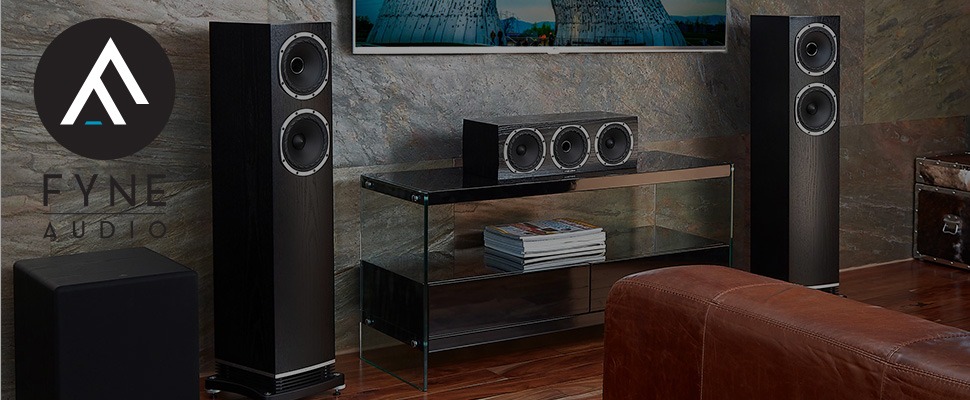 Modern home audio system with two tall black speakers and a central speaker on a sleek glass stand. Background features a stone-textured wall. Fyne Audio logo on the left conveys an elegant, high-end feel.
