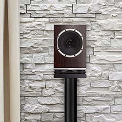 A wooden speaker on a black stand is positioned against a white textured stone wall. The setting is modern and minimalist.