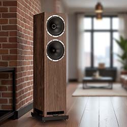 Tall wooden speaker with two circular drivers stands against a brick wall in a modern living room. Soft light and neutral tones create a cosy atmosphere.