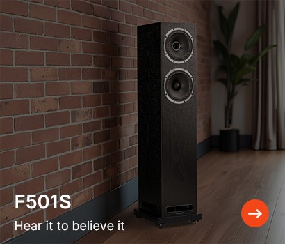 A sleek black speaker with dual woofers stands against a red brick wall on a wooden floor. Text reads "F501S Hear it to believe it," with an arrow icon.