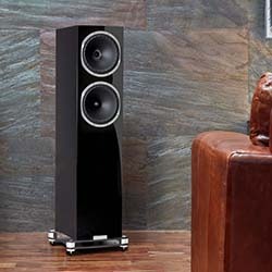 A sleek black floor-standing speaker with two prominent drivers stands on a wooden floor against a textured stone wall, near a brown leather sofa.