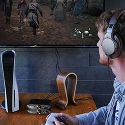 A man with headphones plays a video game on a large screen. A gaming console and VR headset are on a wooden table, creating an immersive setting.