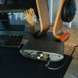 A sleek audio setup on a wooden desk features headphones on a stylish stand, a DAC/amp with a connected cable, and a black keyboard, conveying a modern, tech-savvy vibe.