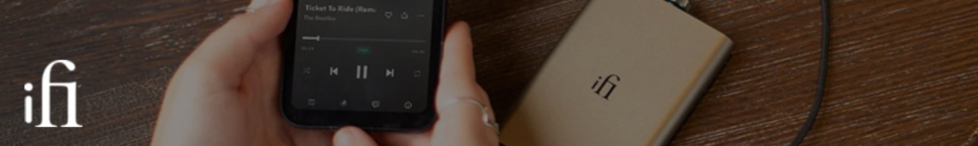 A person holds a smartphone playing "Ticket to Ride" by The Beatles, next to a beige audio device with "ifi" printed on it, on a wooden surface.