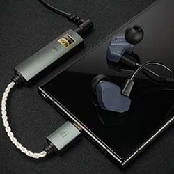 A sleek black smartphone lies on a dark surface, connected to grey earphones and a silver portable amplifier via braided cables.