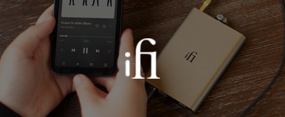 A person holds a smartphone playing "Ticket to Ride" by The Beatles, next to a beige audio device with "ifi" printed on it, on a wooden surface.