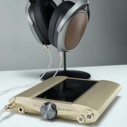A pair of large headphones with metallic grilles on a stand, next to a sleek, gold-coloured audio amplifier with knobs, on a white surface.