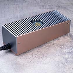 A metallic, rectangular electronic device with vertical black and silver stripes and a power cable on a textured grey surface.