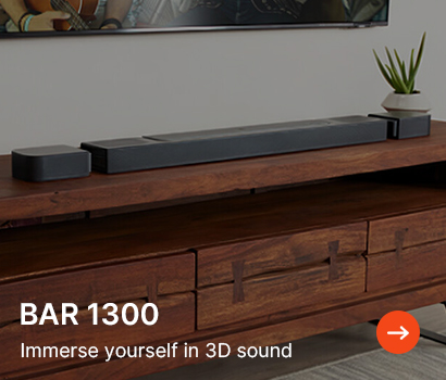 Modern soundbar with two speakers on a wooden console, next to a small plant. Text reads "BAR 1300, Immerse yourself in 3D sound" with an arrow icon.