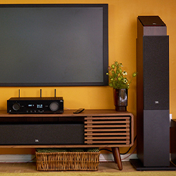 A modern home entertainment setup with a wall-mounted TV on an orange wall, a sleek wooden media console, a black stereo, and a tall speaker.