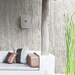 Modern outdoor patio with beige couch, brown and grey pillows against a textured concrete wall. Tall green plants add natural contrast. Serene atmosphere.