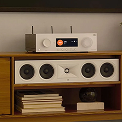 A modern stereo system with a digital display sits atop a wooden shelf. Below it is a sleek speaker with multiple drivers. Books and décor are neatly arranged.