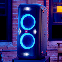 A large black speaker with two glowing blue rings in a dimly lit room, set against a brick wall backdrop, creates a vibrant, energetic atmosphere.